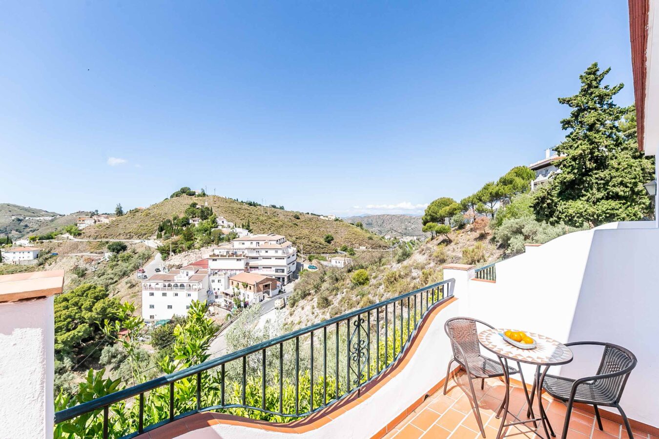 Kullar och vita byggnader. Solbelyst bergsby omgiven av gröna kullar och blå himmel. Charmig vit by belägen på en bergssluttning under blå himmel. Lugnt hörn med pool och solstolar på den spanska landsbygden i söder.