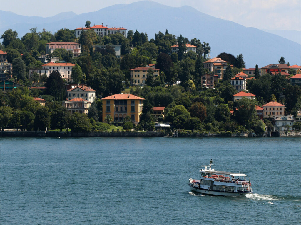 Færge sejler på Lago Maggiore forbi den italienske by Oggebbio med farverige villaer på bjergskråningen.