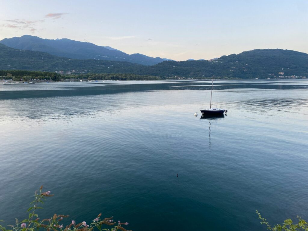 Sejlbåd på stille vand i Lago Maggiore, Italien, med grønne bjerge i baggrunden under en klar himmel.