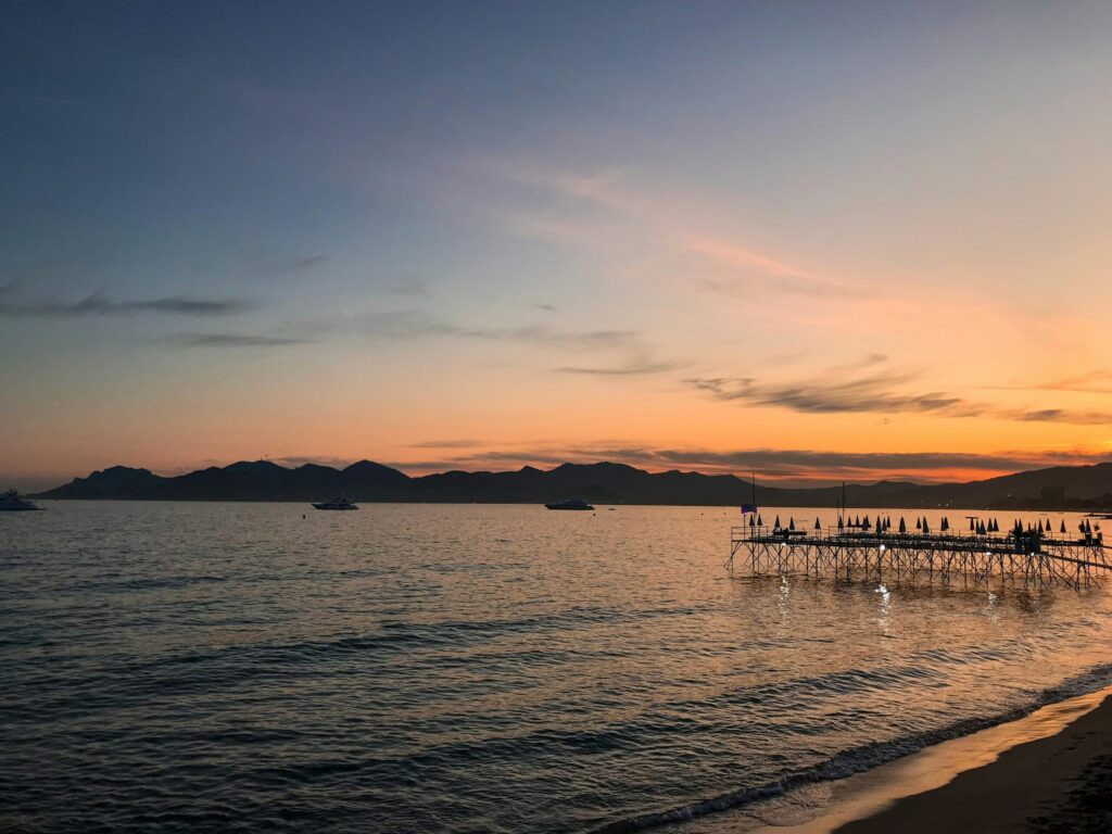 Sunset over a calm sea, with a view of a beach pier with umbrellas, boats on the water, and mountains in the background.