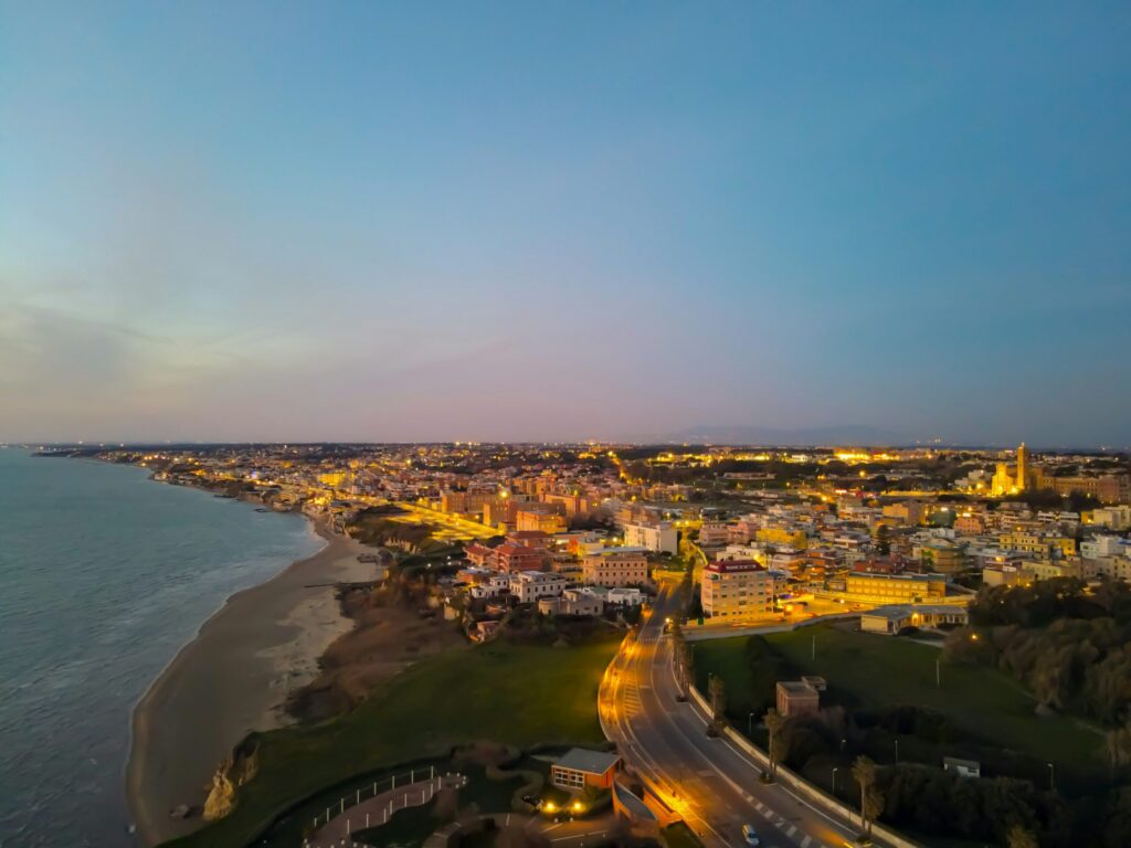 Udsigt over kystbyen ved solnedgang med oplyste gader, bygninger og strandlinje langs havet.