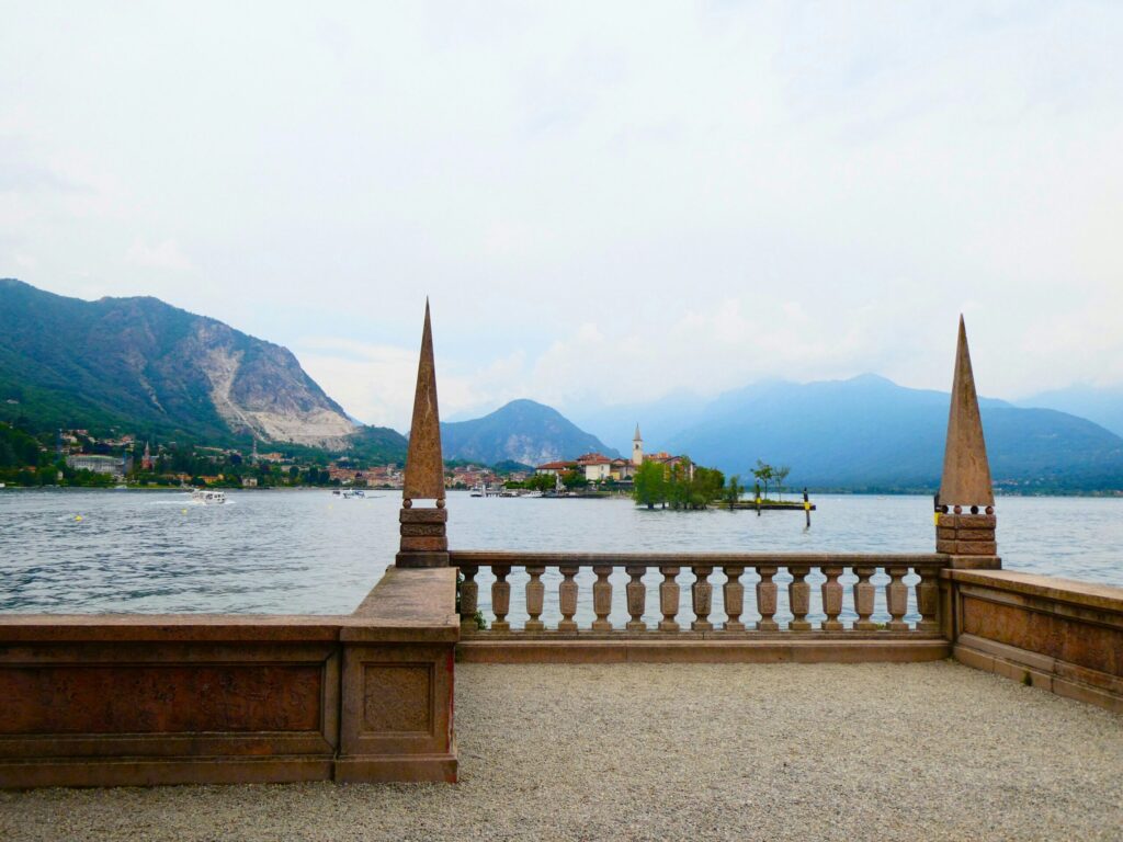 Udsigt fra en stenterrasse mod Isola dei Pescatori i Lago Maggiore, Italien, med bjerge i baggrunden på en overskyet dag.
